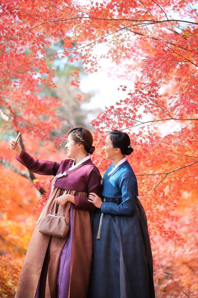 Due donne in abiti tradizionali coreani hanbok si scattano un selfie sotto alberi con foglie autunnali rosse.