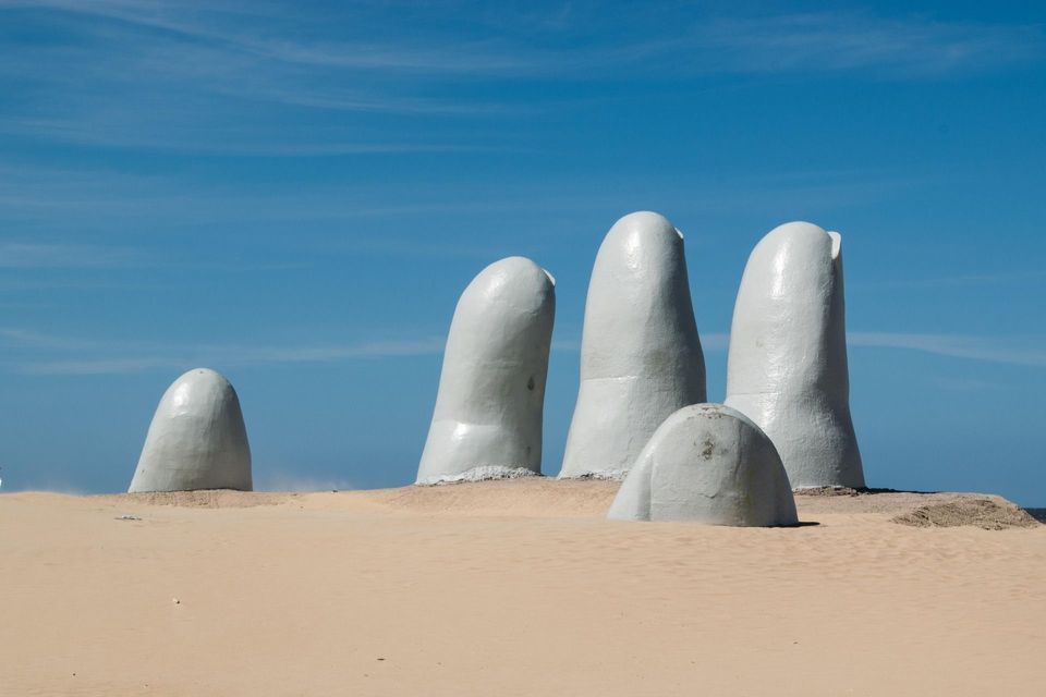 Grande scultura bianca di dita che affiorano dalla sabbia della spiaggia sotto un cielo azzurro.
