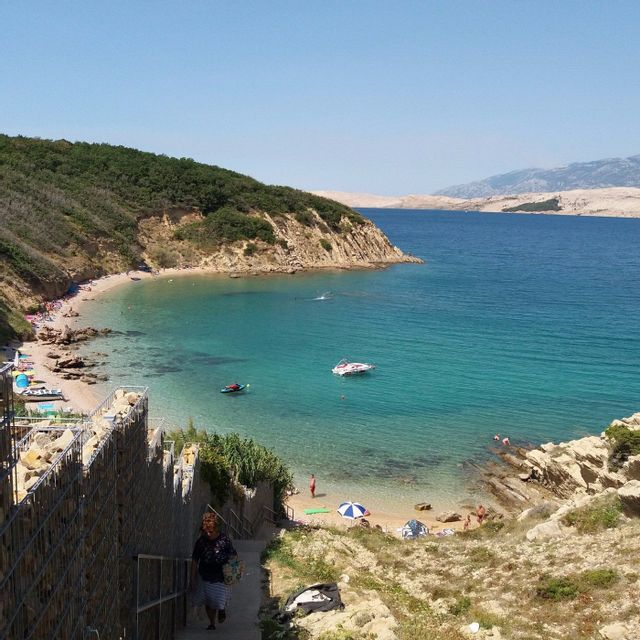 A high-angle view of a sunny coastal cove with turquoise water, a sandy beach with people, and boats floating near the shore.