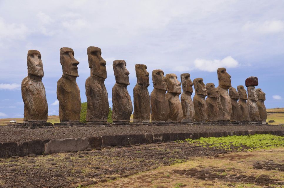 Una lunga fila di grandi statue Moai in pietra che si ergono su una piattaforma in un paesaggio erboso, una delle quali con un copricapo rosso.