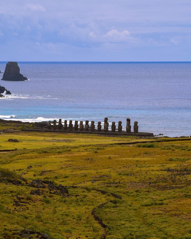 Una fila di grandi statue di pietra si erge su una piattaforma sull'oceano, vista da un vasto campo erboso sotto un cielo nuvoloso.