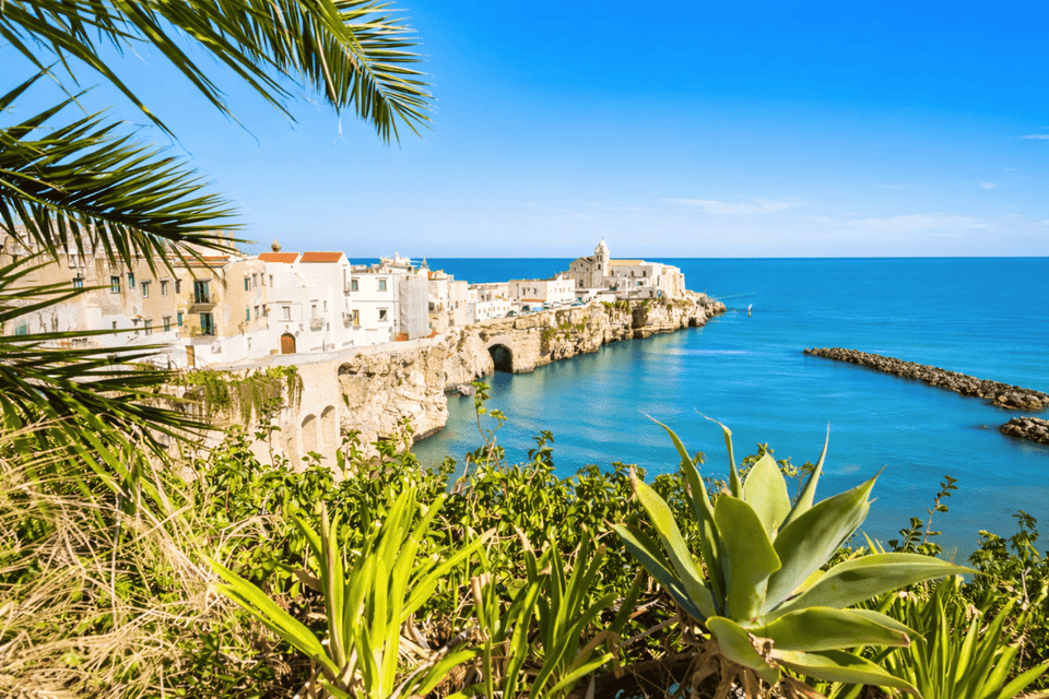 Una città costiera con edifici bianchi, arroccata su una scogliera rocciosa che domina un mare blu calmo, incorniciata da piante verdi e foglie di palma.