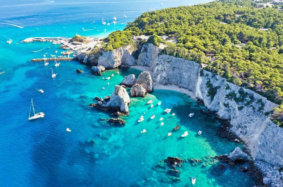 Vista aerea di una costa rocciosa con ripide scogliere bianche e una foresta verde, dove numerose piccole imbarcazioni galleggiano sul mare turchese.