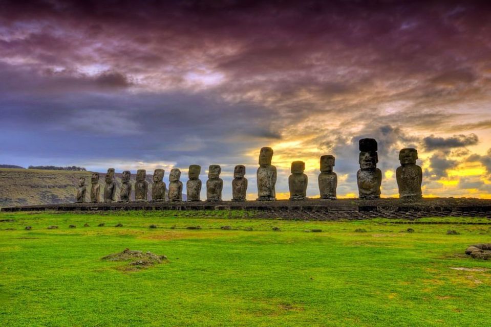 Una fila di grandi statue Moai in pietra, erette su una piattaforma in un campo erboso, sotto un cielo drammatico e nuvoloso al tramonto.