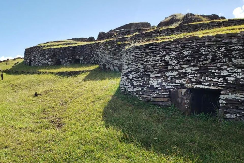 Antiche case in pietra a secco con piccoli ingressi si integrano in un pendio erboso terrazzato, sotto un cielo azzurro.