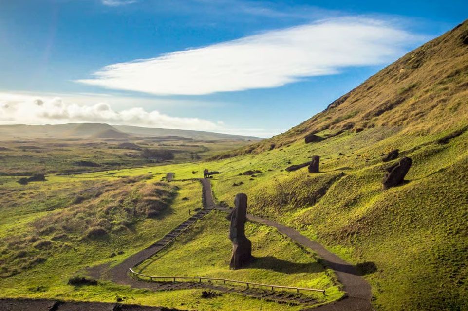 Grandi statue Moai sono sparse su un pendio erboso con un sentiero tortuoso sotto un cielo parzialmente nuvoloso.