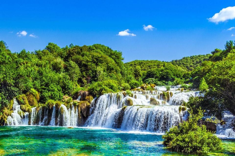 A tiered waterfall cascades into a turquoise river, surrounded by a dense green forest under a bright blue sky.