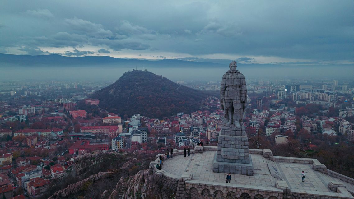 Un grand monument en pierre d'un soldat se dresse sur une colline surplombant une ville tentaculaire avec des montagnes lointaines sous un ciel nuageux.