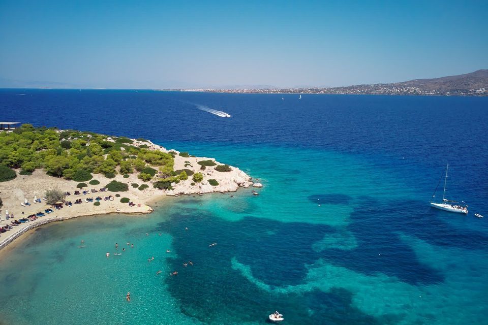 Vista aerea di una piccola penisola con una spiaggia di sabbia, dove le persone nuotano nel mare turchese e una barca a vela è ormeggiata nella baia.