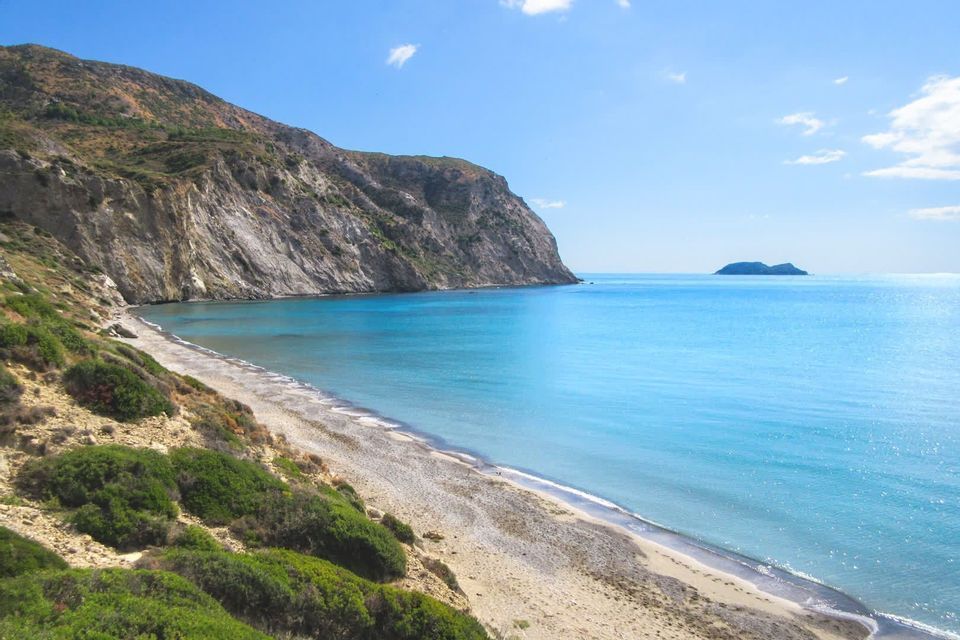 Erhöhter Blick auf einen einsamen Strand mit türkisfarbenem Wasser, das sich am Fuße einer großen Felsklippe unter blauem Himmel erstreckt.