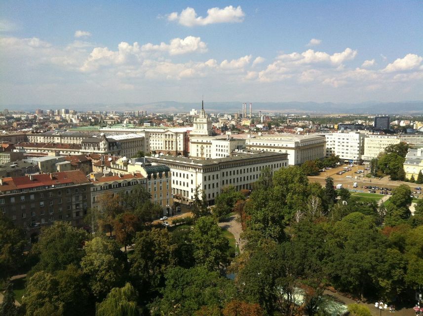 Una vista aerea di una città europea caratterizzata da grandi edifici storici bianchi, un lussureggiante parco verde e montagne lontane sotto un cielo blu.