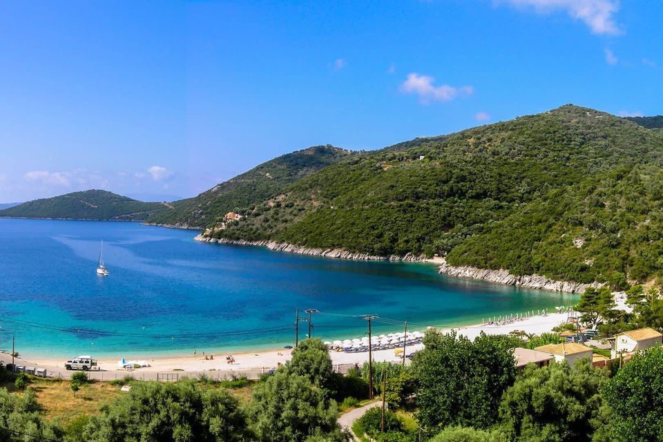 Ein Panoramablick auf einen Sandstrand in einer türkisfarbenen Bucht, umgeben von grünen Hügeln. Ein einzelnes Segelboot schwimmt auf dem ruhigen Wasser.