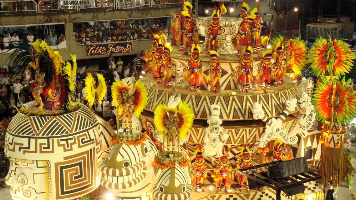 Darsteller in leuchtend gelben und roten Federkostümen tanzen auf einem großen, geschmückten Festwagen während einer nächtlichen Karnevalsparade.