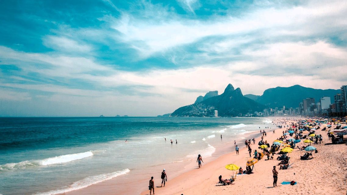 Ein weitläufiger Blick auf einen belebten Strand, an dem Menschen schwimmen und unter Sonnenschirmen entspannen, mit hohen Bergen, die sich hinter einer Küstenstadt erheben.