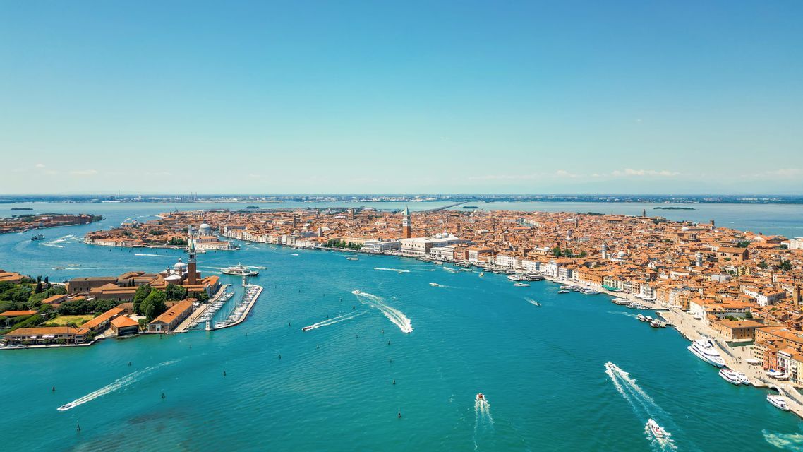 Una vista aerea di Venezia, con i suoi storici edifici dai tetti rossi e le barche che solcano i canali turchesi in una giornata di sole.