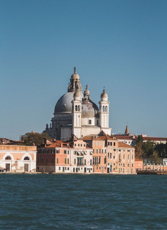 Una grande basilica a cupola bianca si erge sopra edifici colorati sul lungomare sotto un cielo azzurro e limpido.