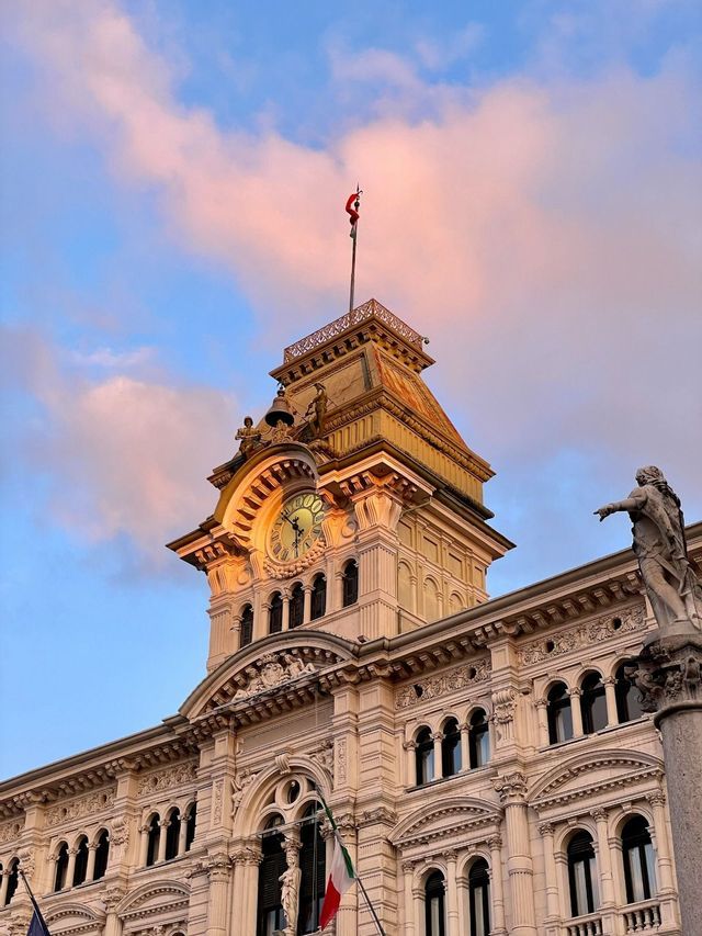 Uno scatto dal basso di un'ornata torre dell'orologio su un edificio storico, contro un cielo con nuvole rosa al tramonto.