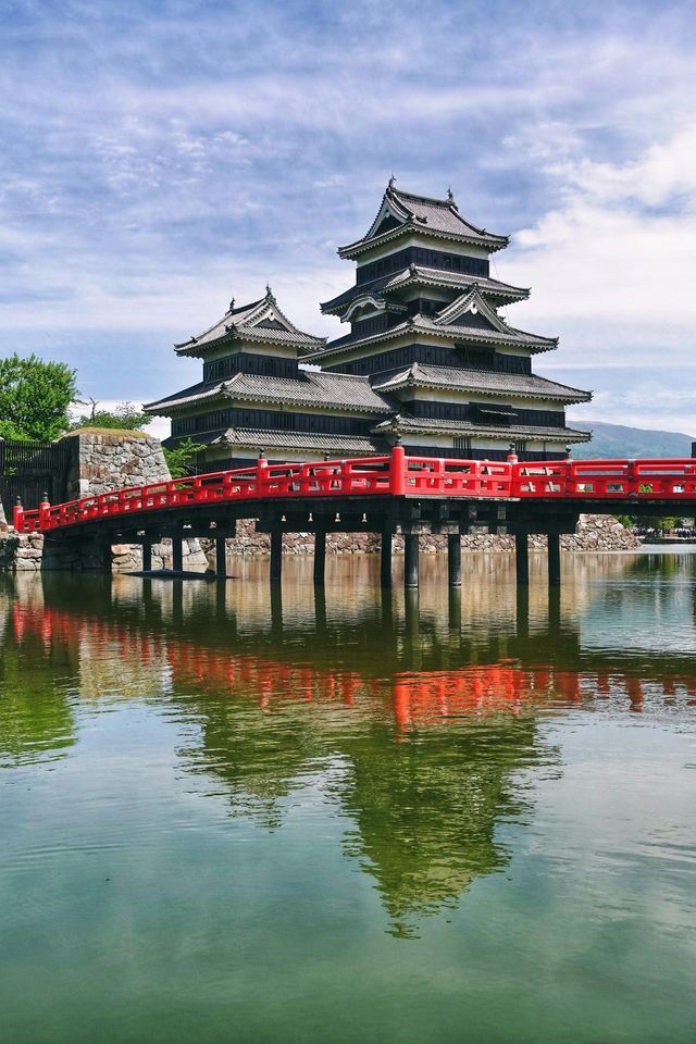 Un castello giapponese tradizionale con un ponte rosso brillante di fronte, entrambi riflessi nelle calme acque di un fossato sotto un cielo nuvoloso.