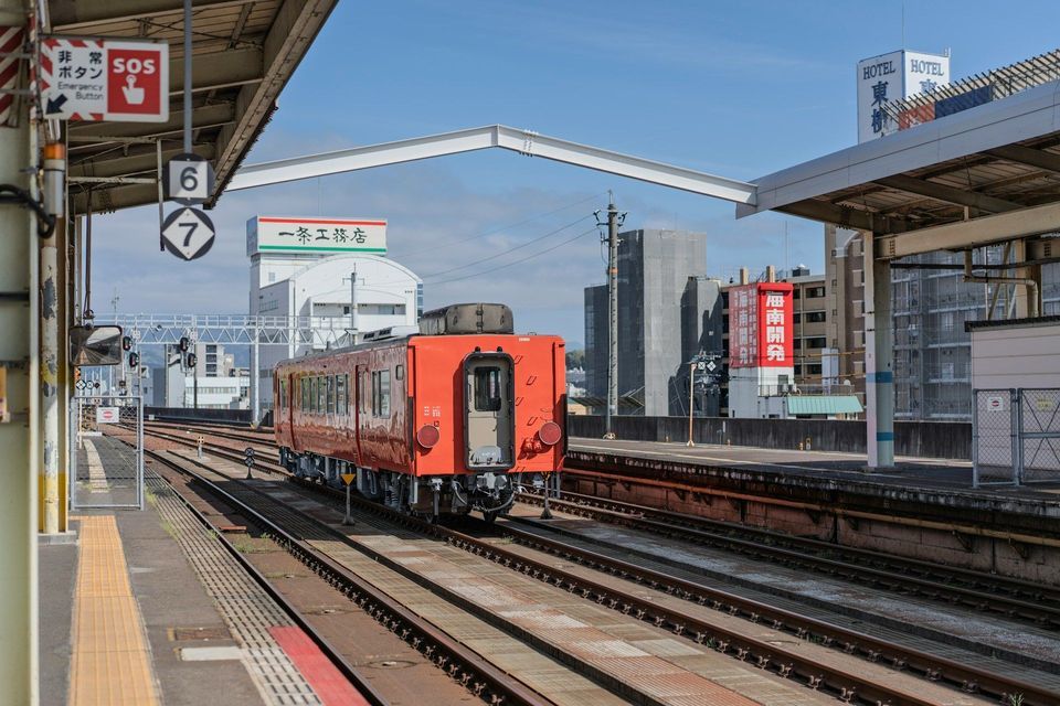 Un treno arancione sui binari su una banchina di stazione all'aperto con edifici cittadini visibili sullo sfondo sotto un cielo azzurro.