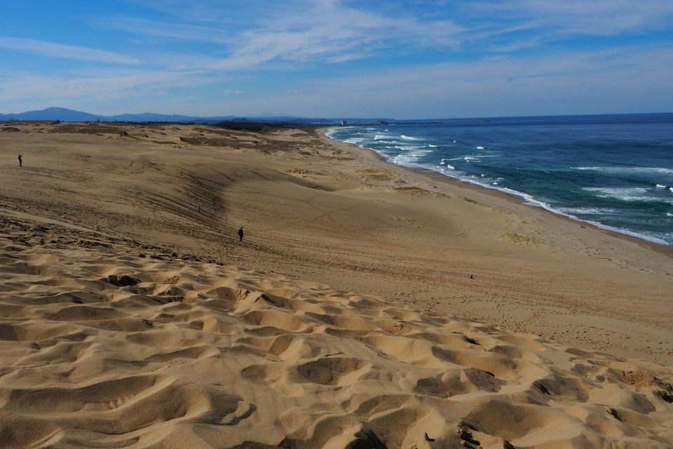 Vaste dune di sabbia che conducono a una costa oceanica blu, con alcune figure distanti che camminano sotto un cielo parzialmente nuvoloso.