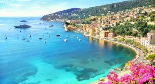 Una città costiera su una collina che domina una baia con acqua turchese e cristallina e molte barche a vela, con fiori rosa in primo piano.