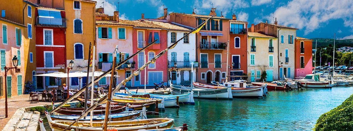 Edifici colorati si affacciano su un porto soleggiato pieno di piccole barche ormeggiate sotto un cielo blu con nuvole bianche.