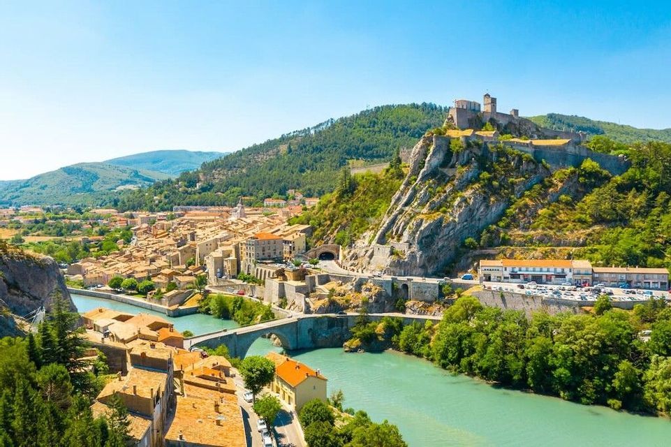 Una cittadella sorge su una rupe rocciosa che domina un borgo con un fiume turchese e un ponte di pietra, circondata da verdi colline.