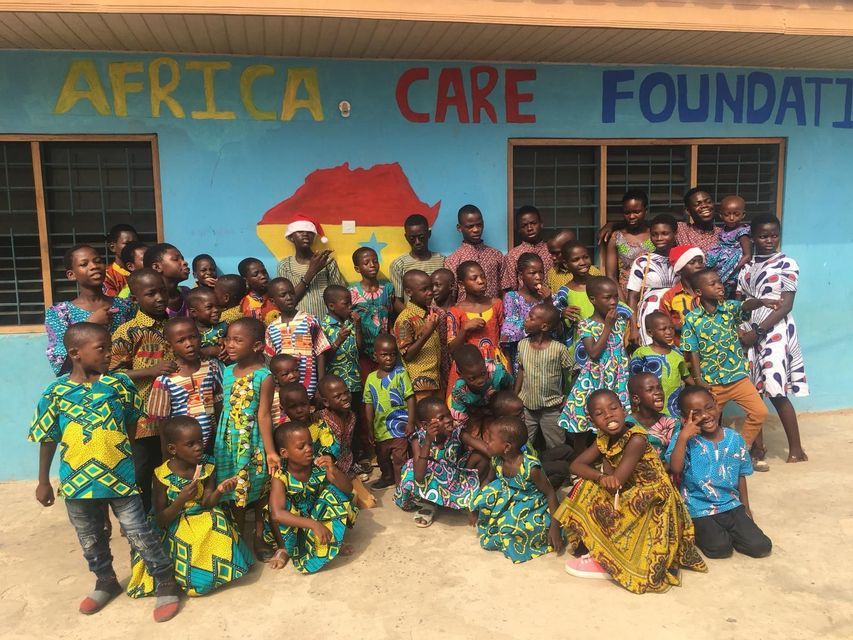 Un folto gruppo di bambini e alcuni adulti con abiti colorati posano per una foto davanti a un edificio blu con scritto 'Africa Care Foundation'.