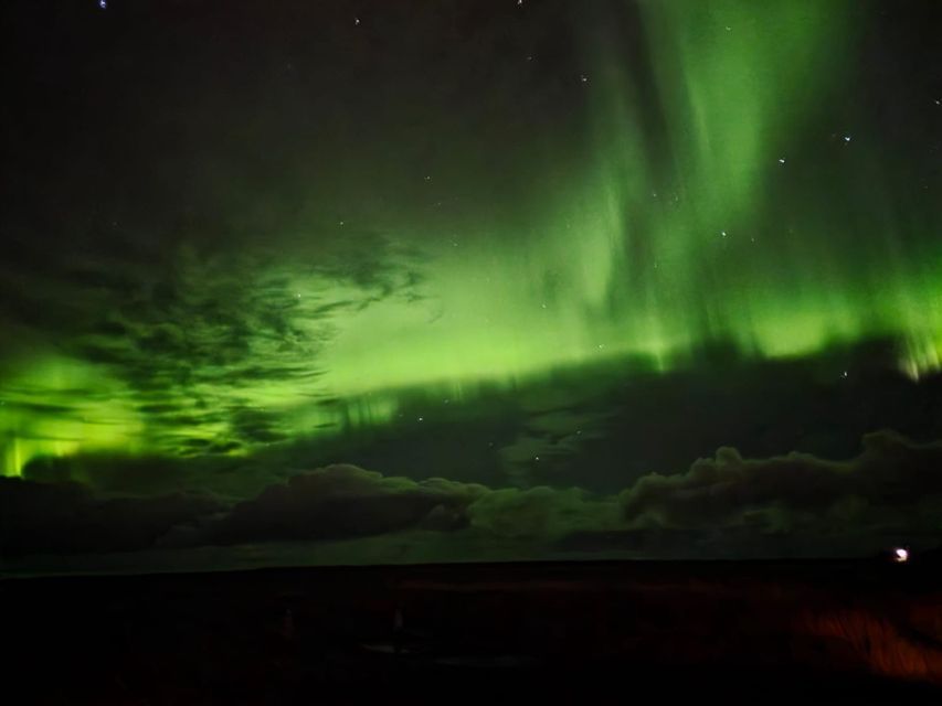 Le luci verdi dell'aurora boreale illuminano un cielo notturno scuro, pieno di stelle e nuvole, sopra un orizzonte buio.