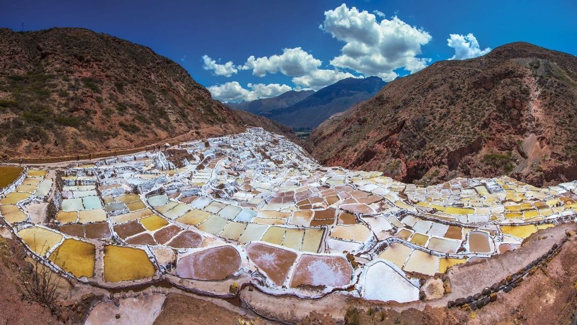Una vista panorámica de salinas en terrazas de varios colores anidadas en un valle de montaña bajo un cielo azul y nublado.