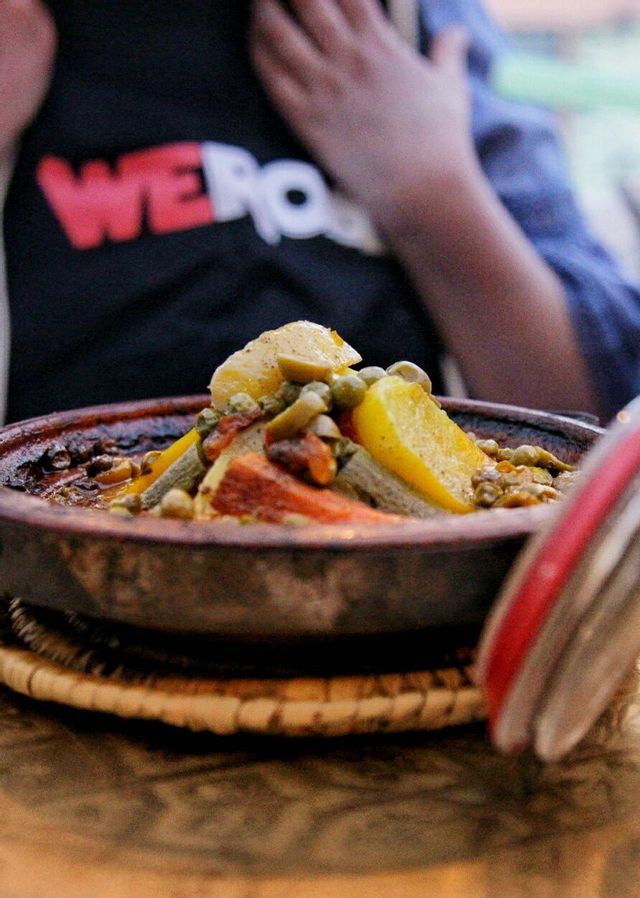 A close-up of a vegetable tagine in a clay pot, with a person wearing a WeRoad t-shirt in the background.