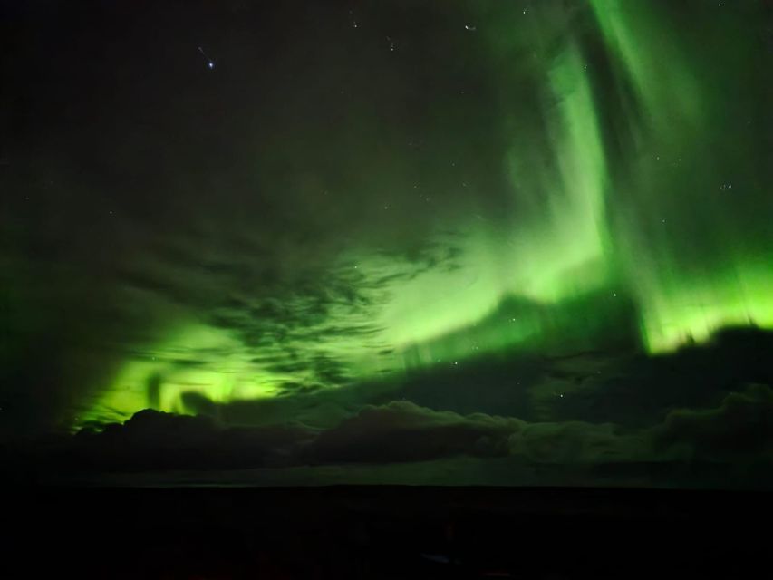 La vivace aurora boreale verde illumina il cielo notturno, serpeggiando tra nuvole e stelle su un paesaggio oscuro.