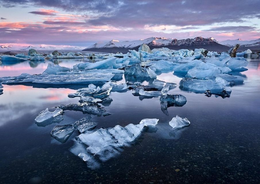 Iceberg e blocchi di ghiaccio galleggiano in una laguna calma al tramonto, con montagne innevate in lontananza sotto un cielo rosa e viola.