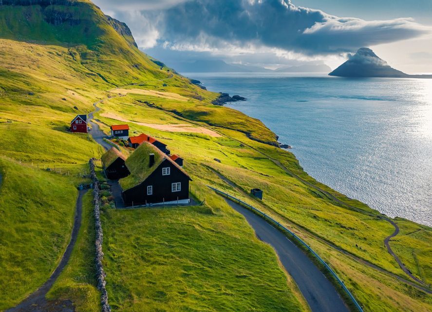 Una vista aerea di case con tetti ricoperti d'erba, adagiate su una rigogliosa scogliera verde a picco sull'oceano sotto un cielo nuvoloso.
