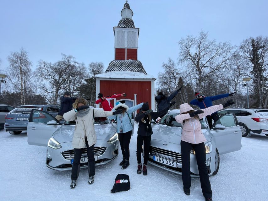 Un grupo de WeRoad posa haciendo el 'dab' junto a dos coches en un paisaje nevado, con un edificio rojo al fondo.