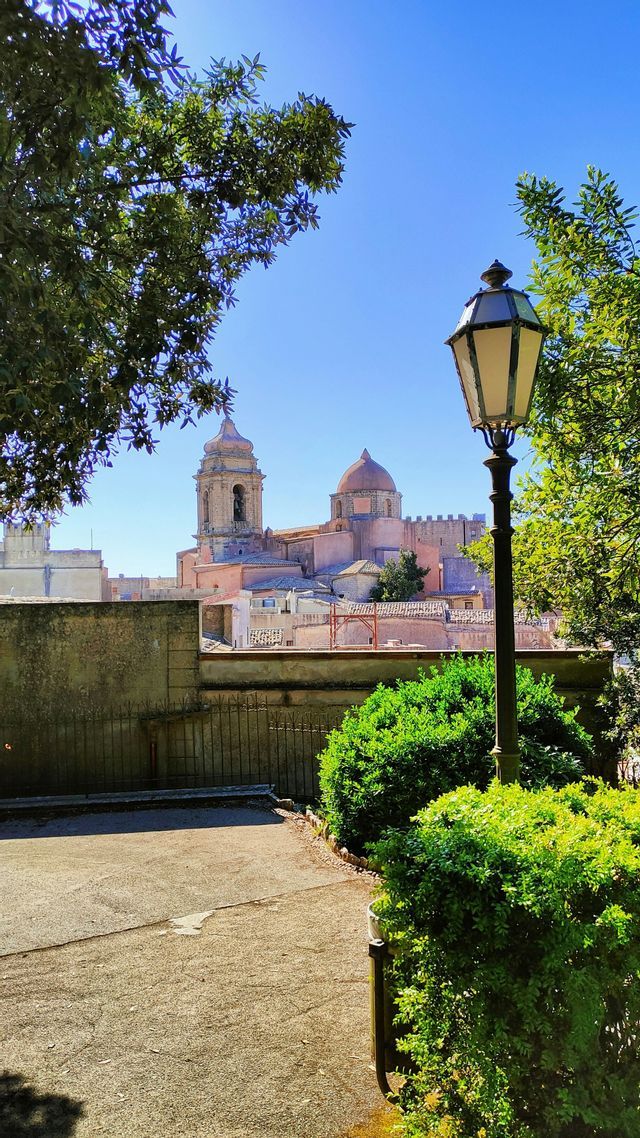 Una vista di una città storica con una cattedrale imponente, incorniciata da alberi verdi e un lampione classico sotto un cielo azzurro.