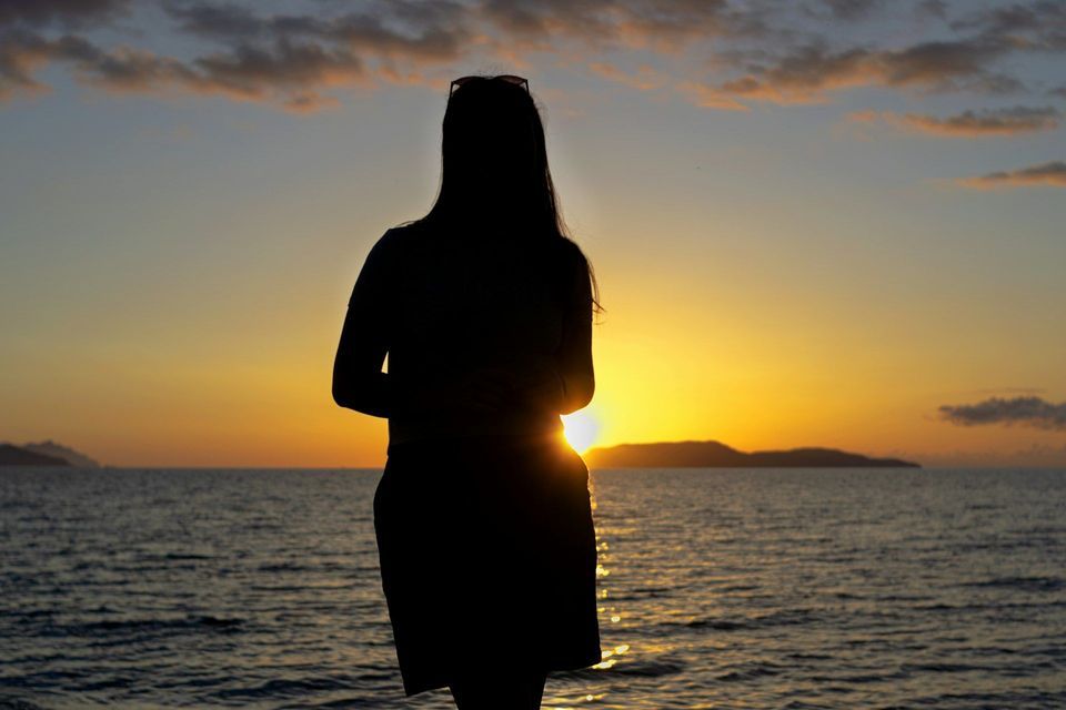 La silhouette di una persona con capelli lunghi che guarda un tramonto dorato sul mare, con isole lontane all'orizzonte.