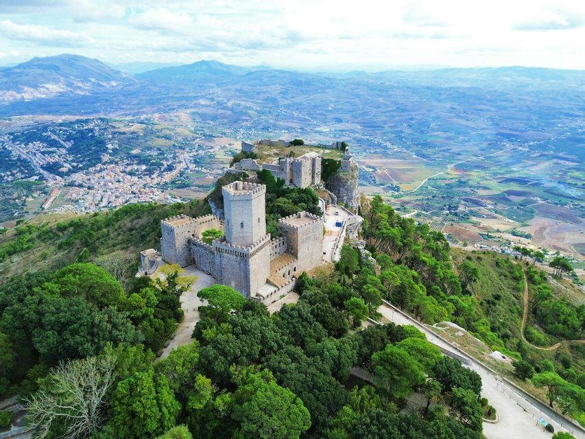 Una vista aerea di un castello storico in pietra, situato su una montagna boscosa, che domina un'ampia valle e una città lontana.