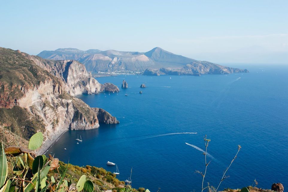 Una vista dall'alto di una costa aspra e rocciosa che si fonde con un mare blu intenso, con isole vulcaniche all'orizzonte e diverse imbarcazioni sull'acqua.