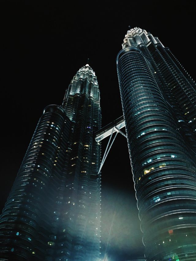 Una vista en ángulo bajo de dos rascacielos modernos con un puente aéreo que los conecta, iluminados contra un cielo nocturno oscuro.