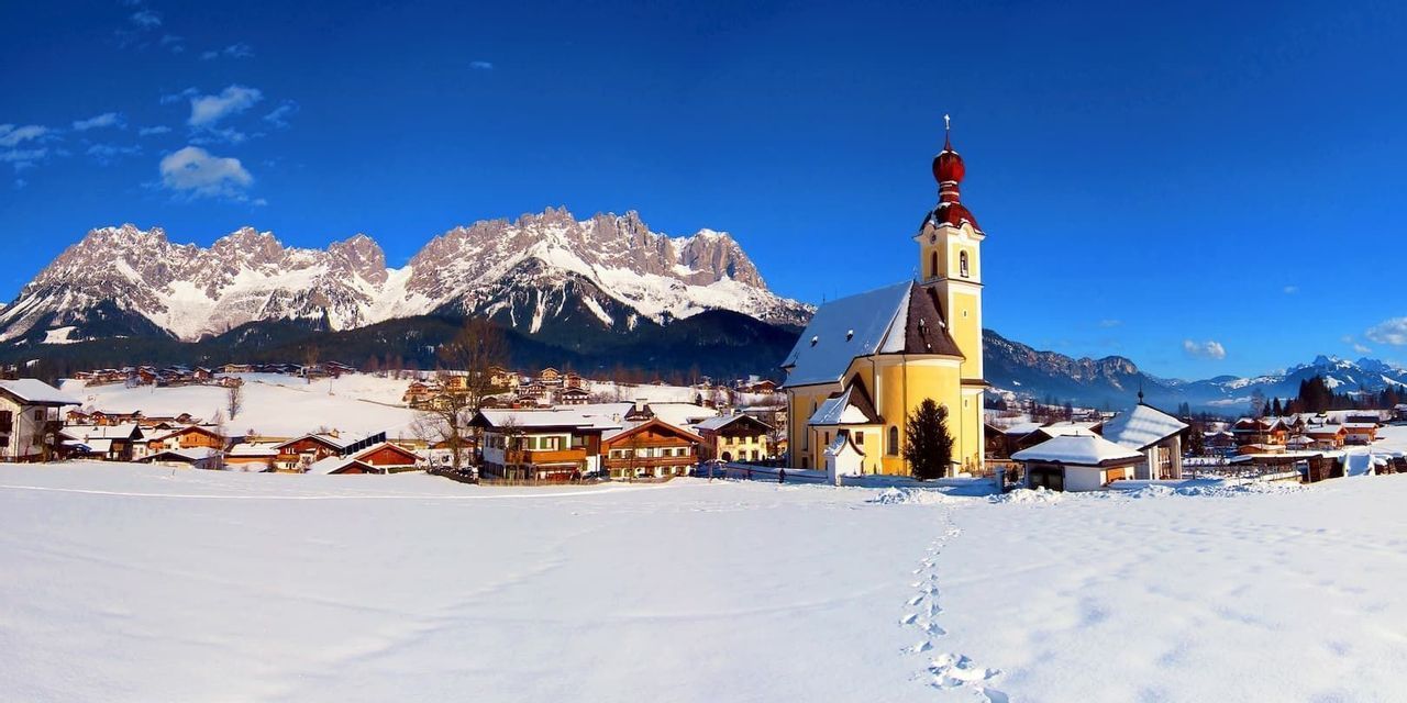 Eine gelbe Kirche in einem schneebedeckten Dorf mit Fußspuren im Vordergrund, vor einer Kulisse von Bergen unter einem klaren blauen Himmel.