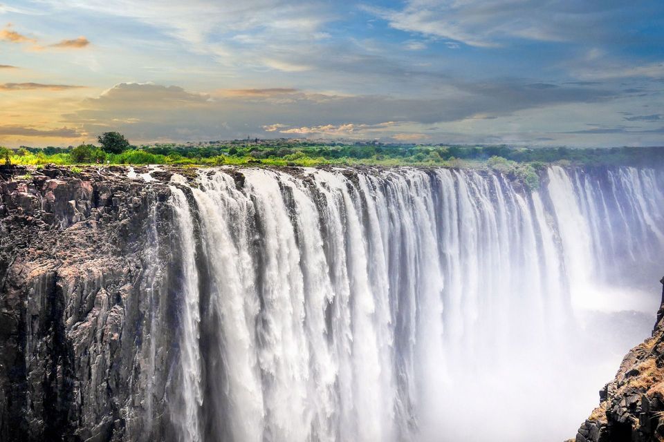 A massive waterfall rushes over a wide, rocky cliff edge, with green vegetation visible in the background under a blue and cloudy sky.