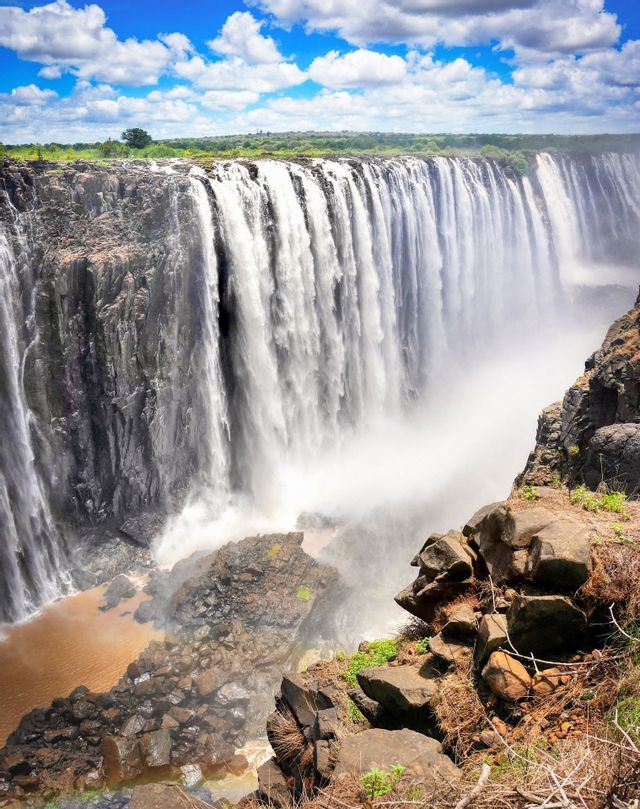 A wide waterfall cascades down a rocky gorge, sending up mist into a cloudy blue sky.