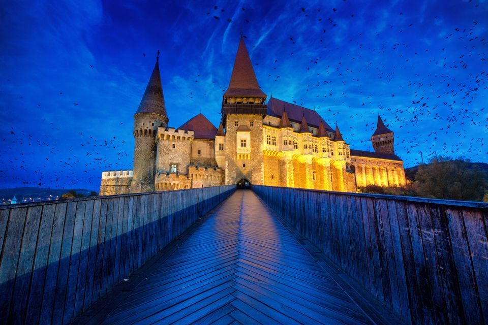 Un largo puente de madera conduce a un gran castillo de piedra iluminado al atardecer, con una bandada de pájaros en el cielo azul profundo.