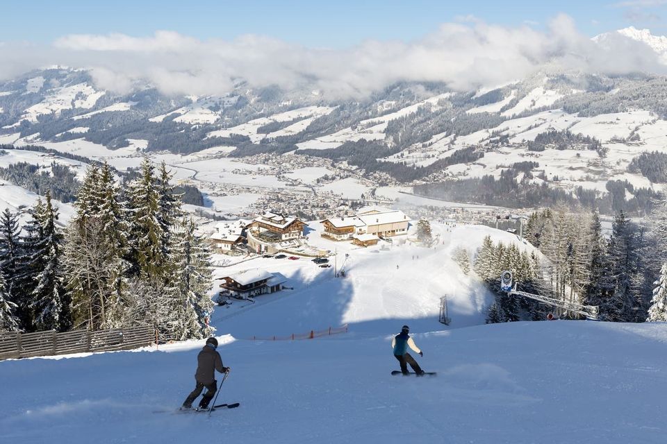 Ein Skifahrer und ein Snowboarder von einer WeRoad Gruppenreise fahren einen sonnigen Hang oberhalb eines schneebedeckten Bergdorfs hinab.