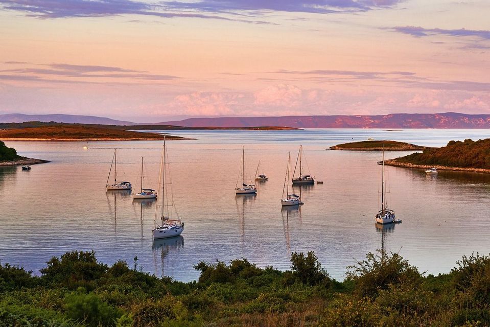Several sailboats are anchored in a calm bay surrounded by green hills under a colorful sunset sky.