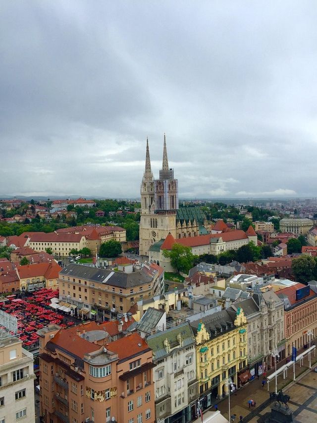 A high-angle view of a cityscape featuring a large, twin-spired cathedral surrounded by buildings with red roofs under a cloudy sky.