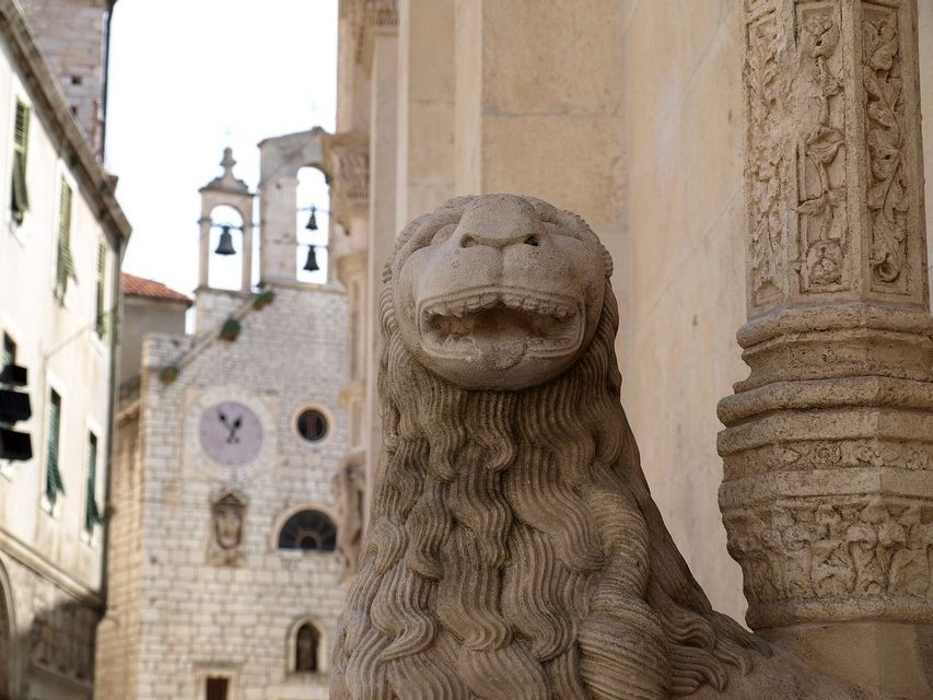 A carved stone lion statue with an open mouth stands guard next to a pillar, with a historic stone clock tower in the background.