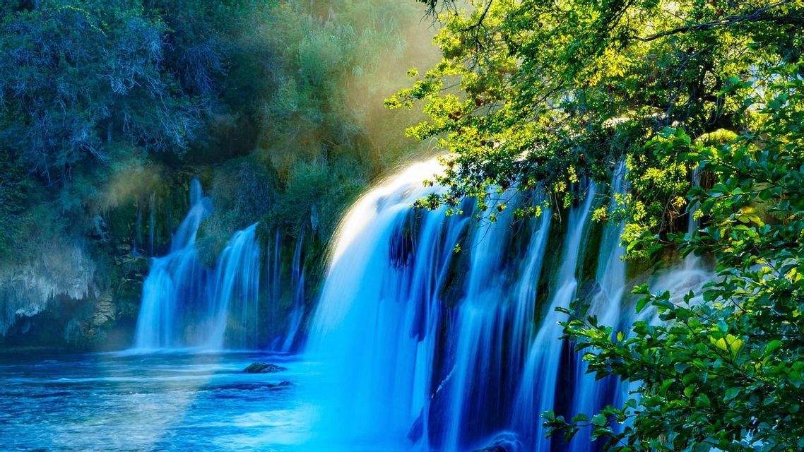 A cascading waterfall with blue, motion-blurred water flows through a lush green forest, with sunlight filtering through the tree branches.