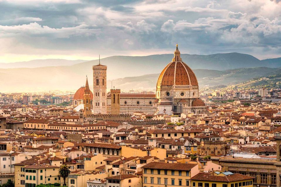 Un extenso paisaje urbano de tejados de terracota con una destacada catedral de cúpula y torre, situado frente a montañas distantes bajo un cielo nublado.